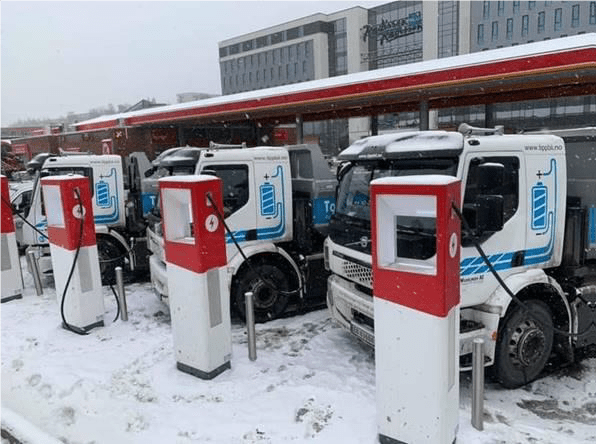 Elektriske lastebiler parkert ved ladestasjoner i et snødekt område nær en moderne bygning.
