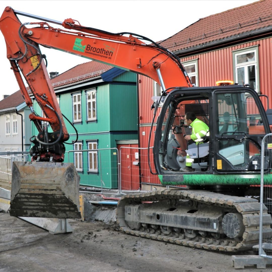 En gravemaskin med en person inne opererer på en byggeplass i en gate, med fargerike hus i bakgrunnen.