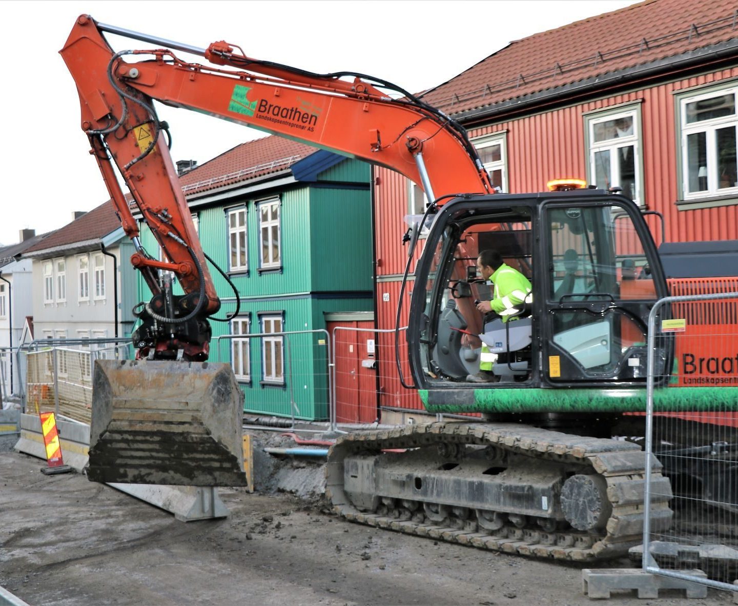 En gravemaskin med en person inne opererer på en byggeplass i en gate, med fargerike hus i bakgrunnen.