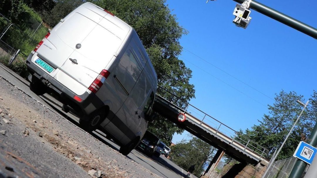 Hvit varebil kjører på en vei mot en bro, med et trafikkkamera over hodet og klar himmel i bakgrunnen.