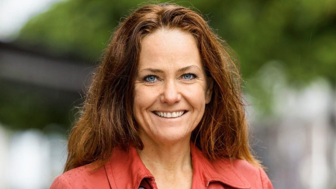 En kvinne med langt rødt hår og blå øyne smiler varmt, iført en rød jakke som står i vakker kontrast til de frodige grønne trærne i den uskarpe bakgrunnen – et perfekt portrett inspirert av klimainvesteringer, som gjenspeiler naturens innvirkning på vår verden.