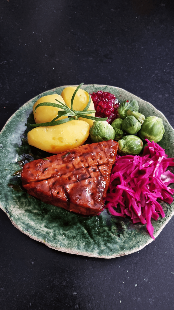En festlig tallerken med grillet tempeh, gule poteter, rosenkål, rødkål og tyttebærsaus på et grønt keramikkfat – perfekt til juleretter uten kjøtt.