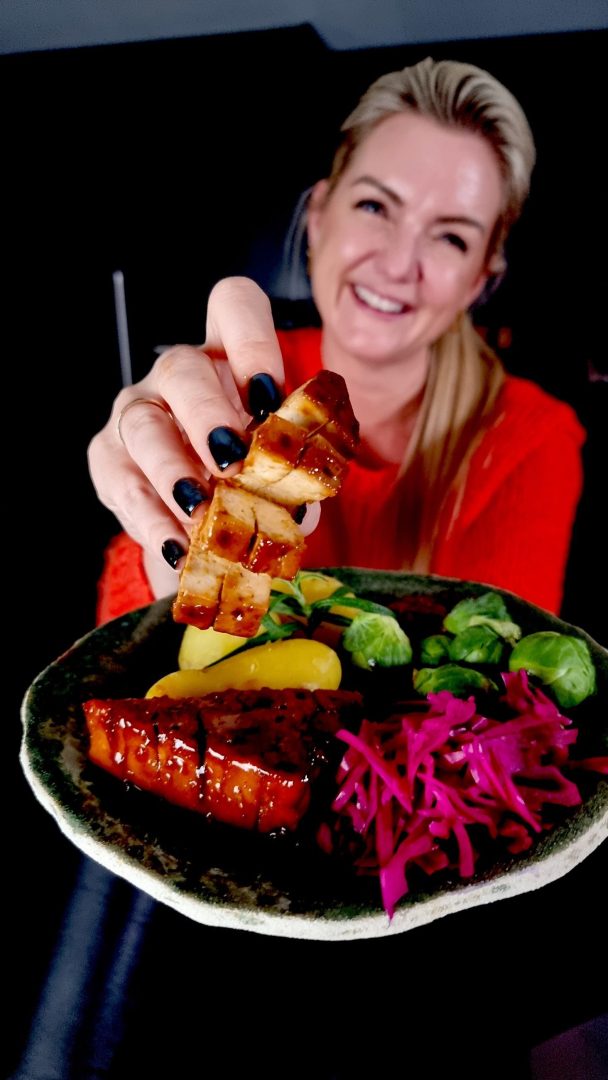 En person som smiler til kameraet og stolt presenterer en tallerken med glasert tofu, grønnsaker i skiver og syltet kål – en innbydende versjon av juleretter uten kjøtt.