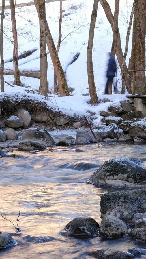 En uskarp figur går forsiktig langs en snødekt sti ved siden av en rennende bekk med steiner, omgitt av nakne trær – en scene som kan passere som et Oslotips-postkort som fanger vinterens rolige skjønnhet.