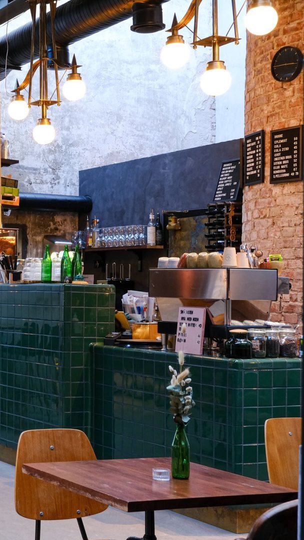 Kaféinteriør i industriell stil med trebord, en grønn flislagt disk, en espressomaskin og hengende lys som fremhever de synlige murveggene. En liten grønn flaske med blomster står på et bord, og gir et sjarmerende preg midt i denne Oslotips-perlen.