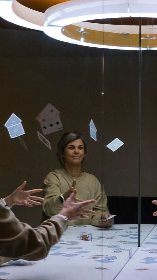 Personen sitter ved bordet og kaster spillekort opp i luften med bekymringsløs oppgivelse. Refleksjonen deres i speilet bak dem fanger hvert gledesøyeblikk, som minner om en scene rett ut av et Oslotips-trylleshow.