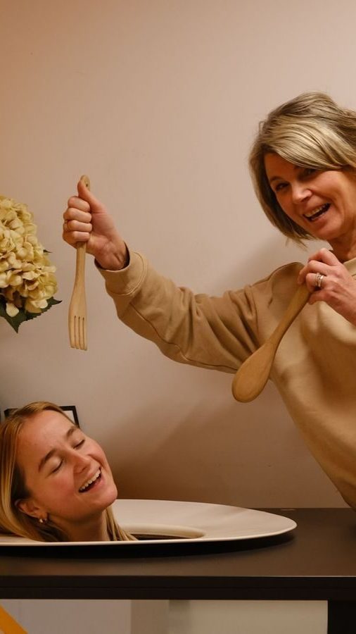 En kvinne holder lekent en gaffel og skje over hodet til en annen person, som dukker opp gjennom bordet i en Oslotips-inspirert setting. Smilene deres er smittsomme mens de deler et øyeblikk med lunefull glede.