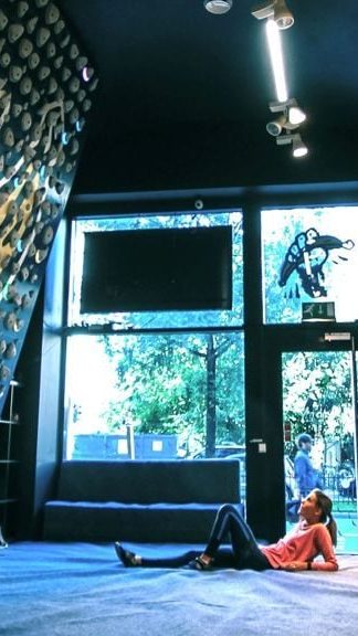 Person som sitter på gulvet i et innendørs klatretreningsstudio og ser opp på klatregrep på en høy vegg. Naturlig lys strømmer gjennom store vinduer, og skaper en rolig og fokusert atmosfære – et ideelt sted for noen Oslotips for å forbedre klatreferdighetene dine.