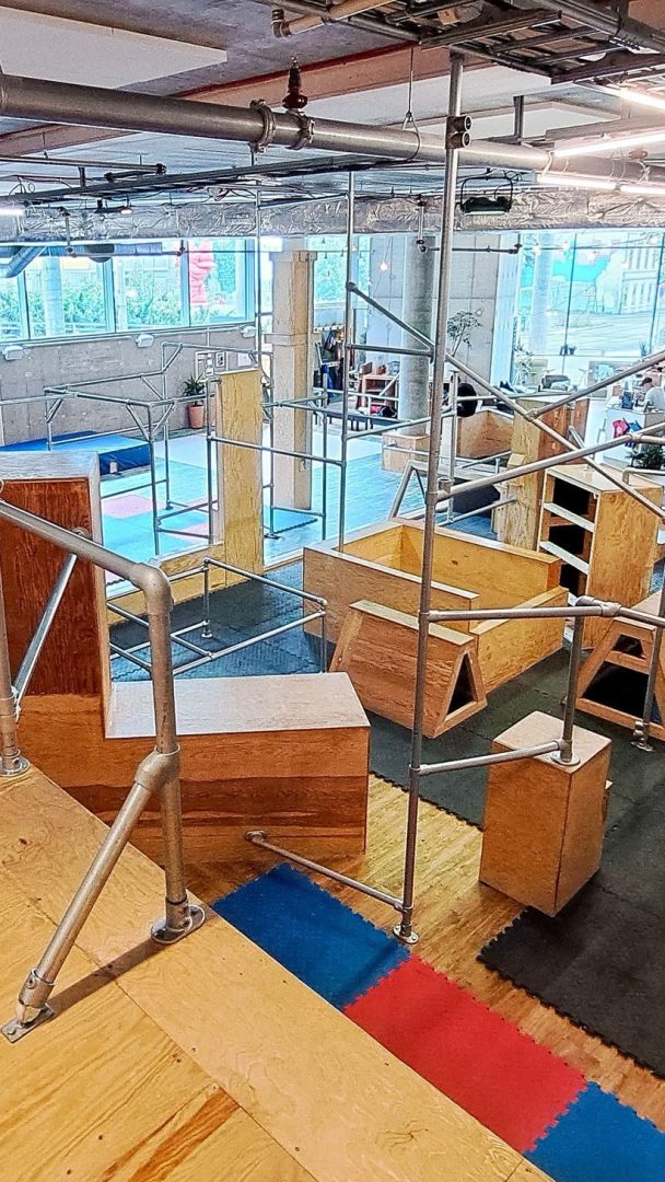 Oslotips: Oppdag et innendørs parkour-treningsstudio med tre- og metallhindringer, inkludert plattformer, barer og ramper på polstret gulv. Store vinduer oversvømmer plassen med naturlig lys, noe som forbedrer treningsopplevelsen din.