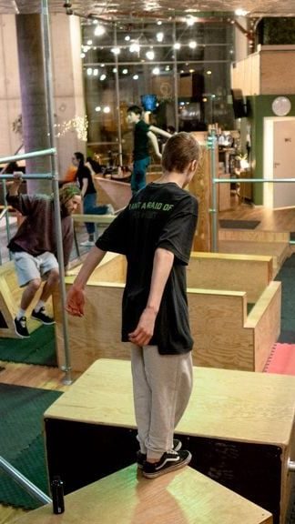 Person som balanserer på en treplattform i et innendørs parkour-gym, med andre i bakgrunnen som øver på hindringer. For en ekte Oslotips-opplevelse tilbyr dette stedet både utfordring og spenning for alle parkour-entusiaster som ønsker å skjerpe ferdighetene sine.