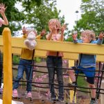 Fire barn leker på en trebro på en lokal lekeplasser. Ett barn holder en teddybjørn. Trær og et lysbilde er synlige i bakgrunnen.