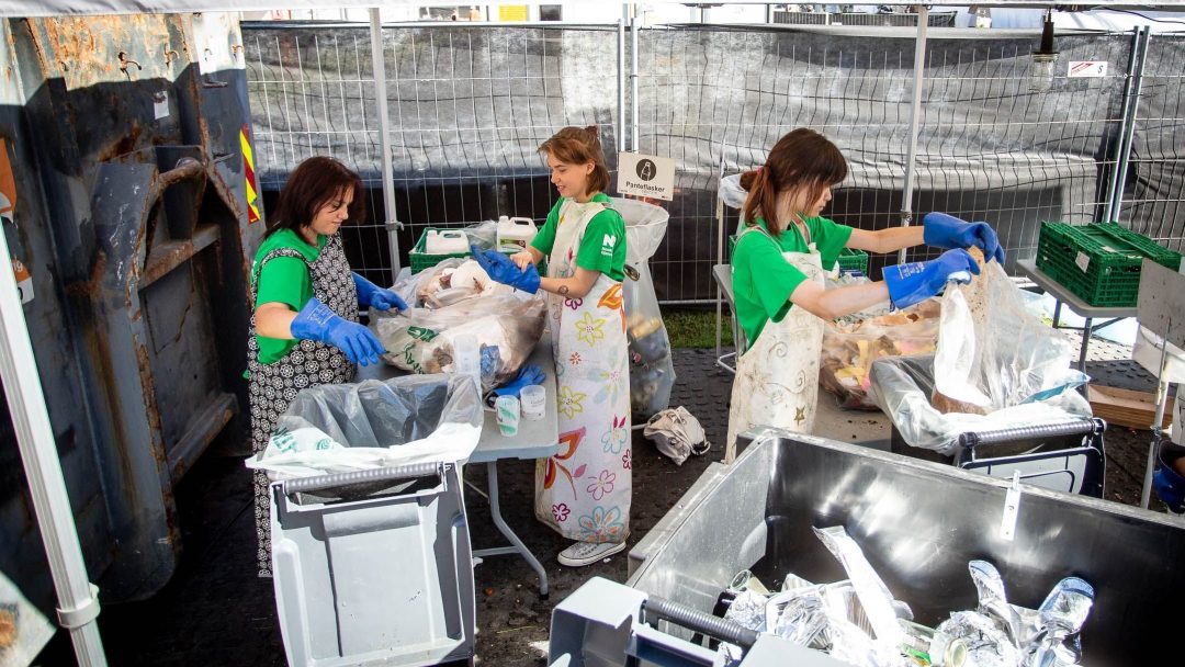 Tre personer i forkle og hansker sorterer avfall i søppelkasser på en utendørs gjenvinningsstasjon.