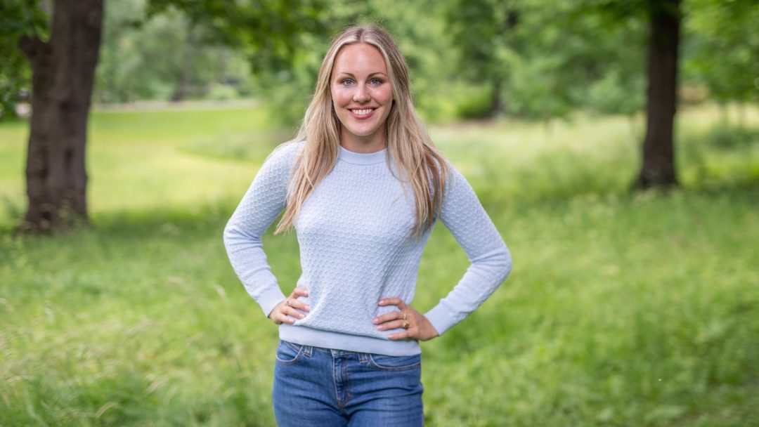 En kvinne iført en lyseblå genser og jeans står i en grønn park med trær, og legemliggjør ånden til Klimapilotene.