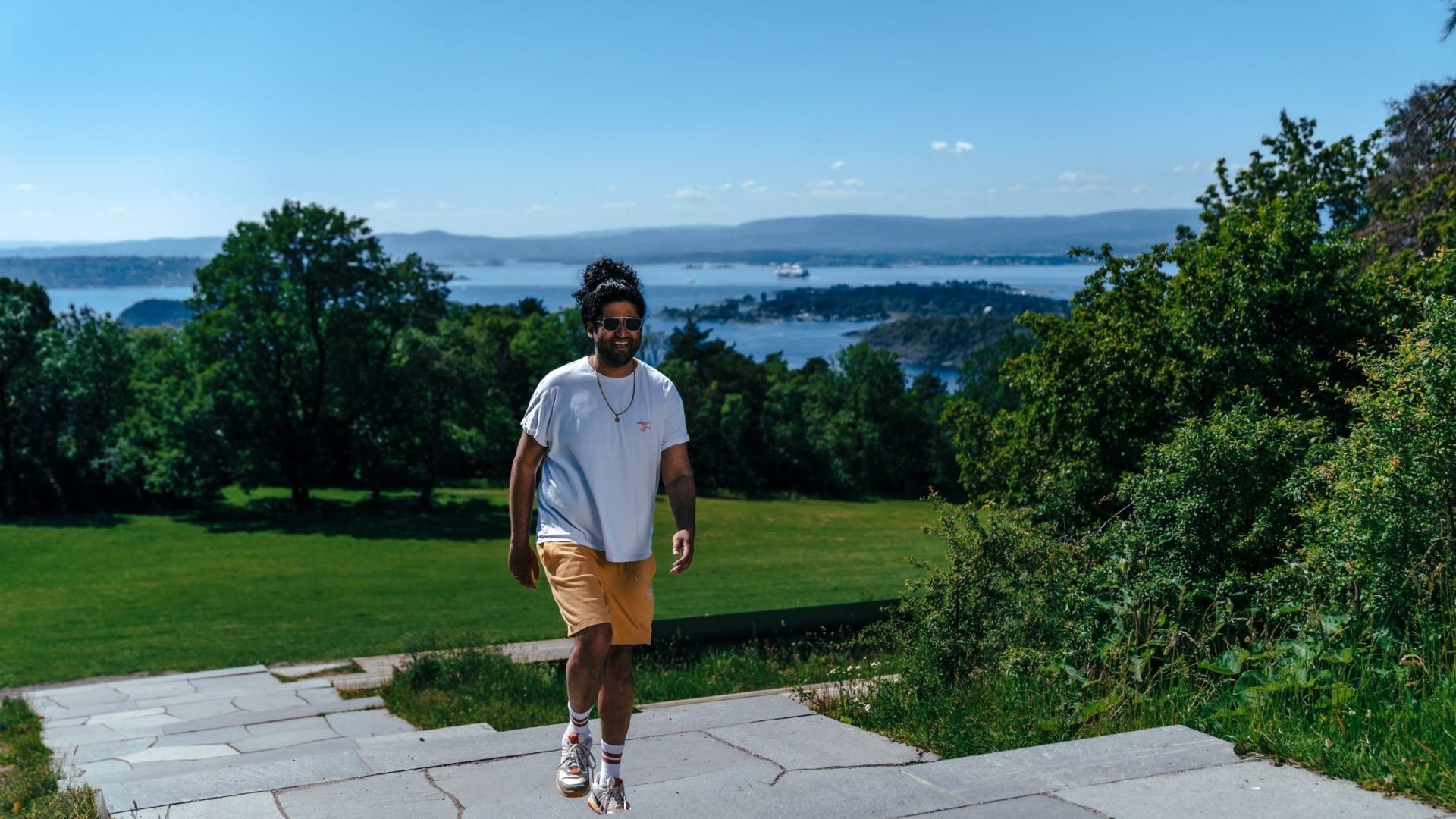 En person i hvit skjorte og gul shorts nyter gåturen langs en steinsti, med naturskjønn utsikt over bukten, frodig grønt gress og trær i bakgrunnen.