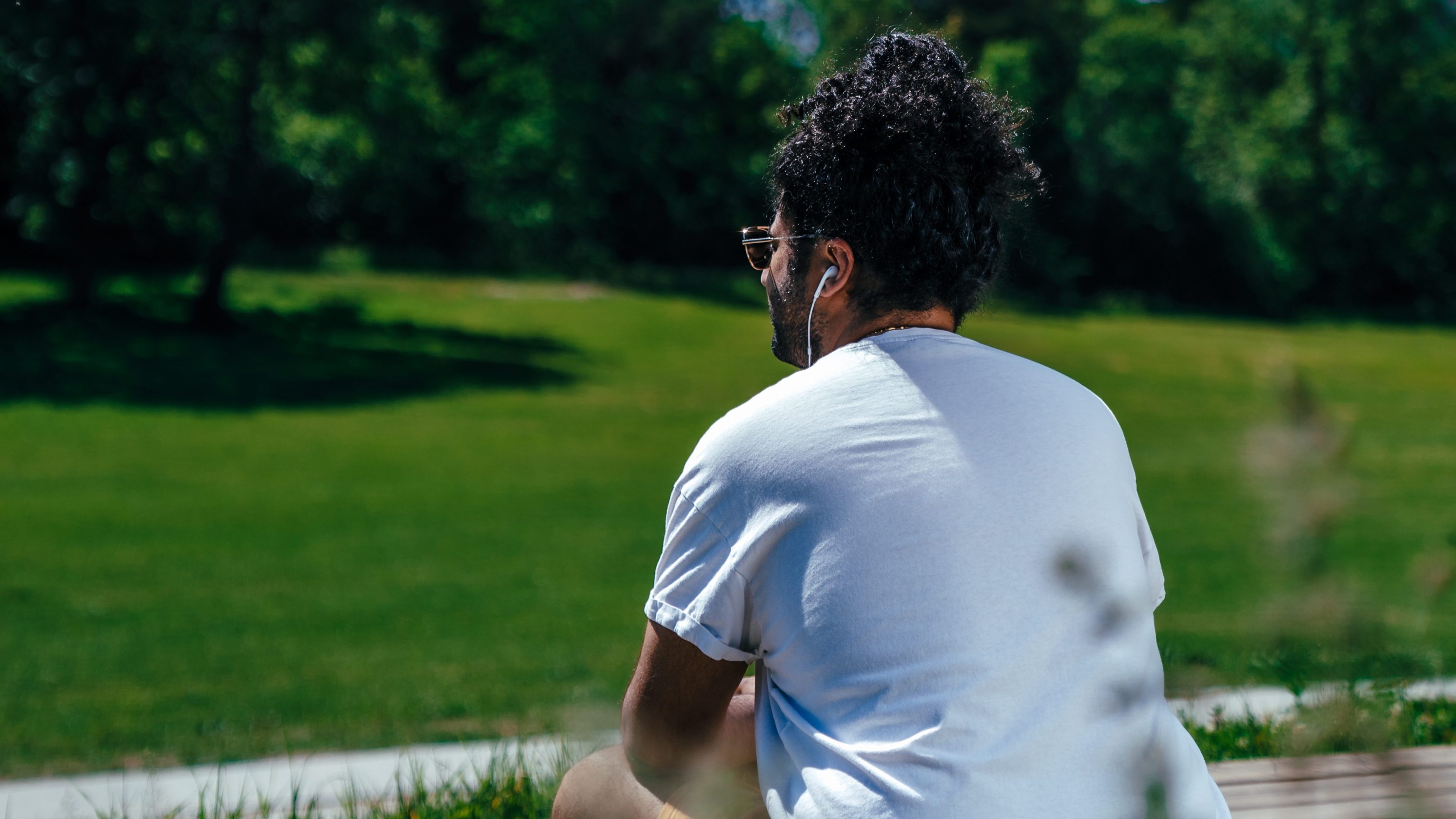 En person med krøllete hår sitter utendørs, nyter en gåturen i en hvit T-skjorte og øretelefoner, vendt mot et gresskledd område med trær.