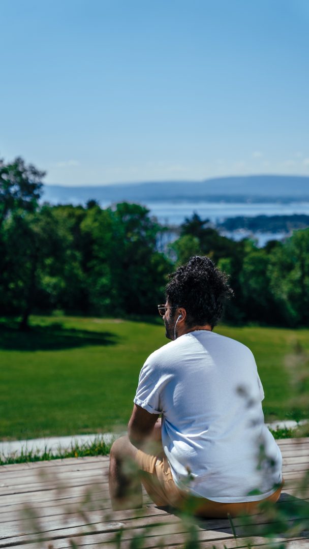 En person i hvit skjorte sitter på et tredekk og hviler etter den forfriskende gåturen, med utsikt over et frodig grønt landskap med en glitrende vannmasse i det fjerne under den klare blå himmelen.