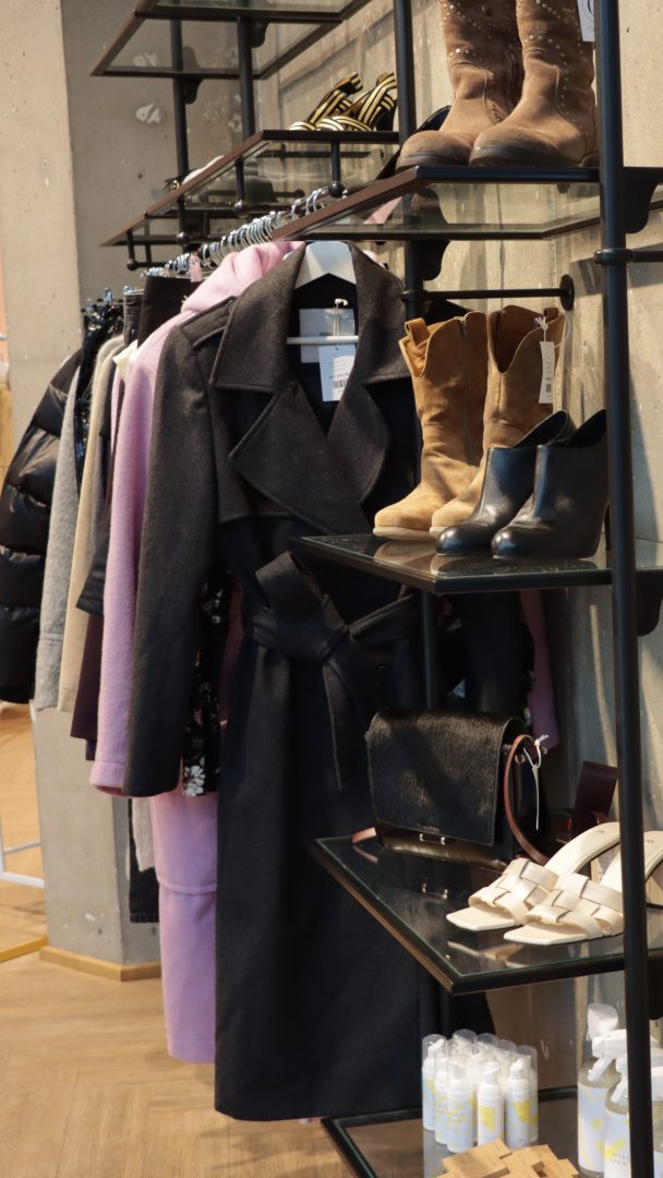 Bruktbutikker-displayet har kåper, sko og vesker i hyllene. Kremfargede sandaler og en svart veske hviler på nederste hylle, med brune støvler og svarte sko over. Kåper henger elegant på et stativ til siden, og viser brukt stil med raffinement.