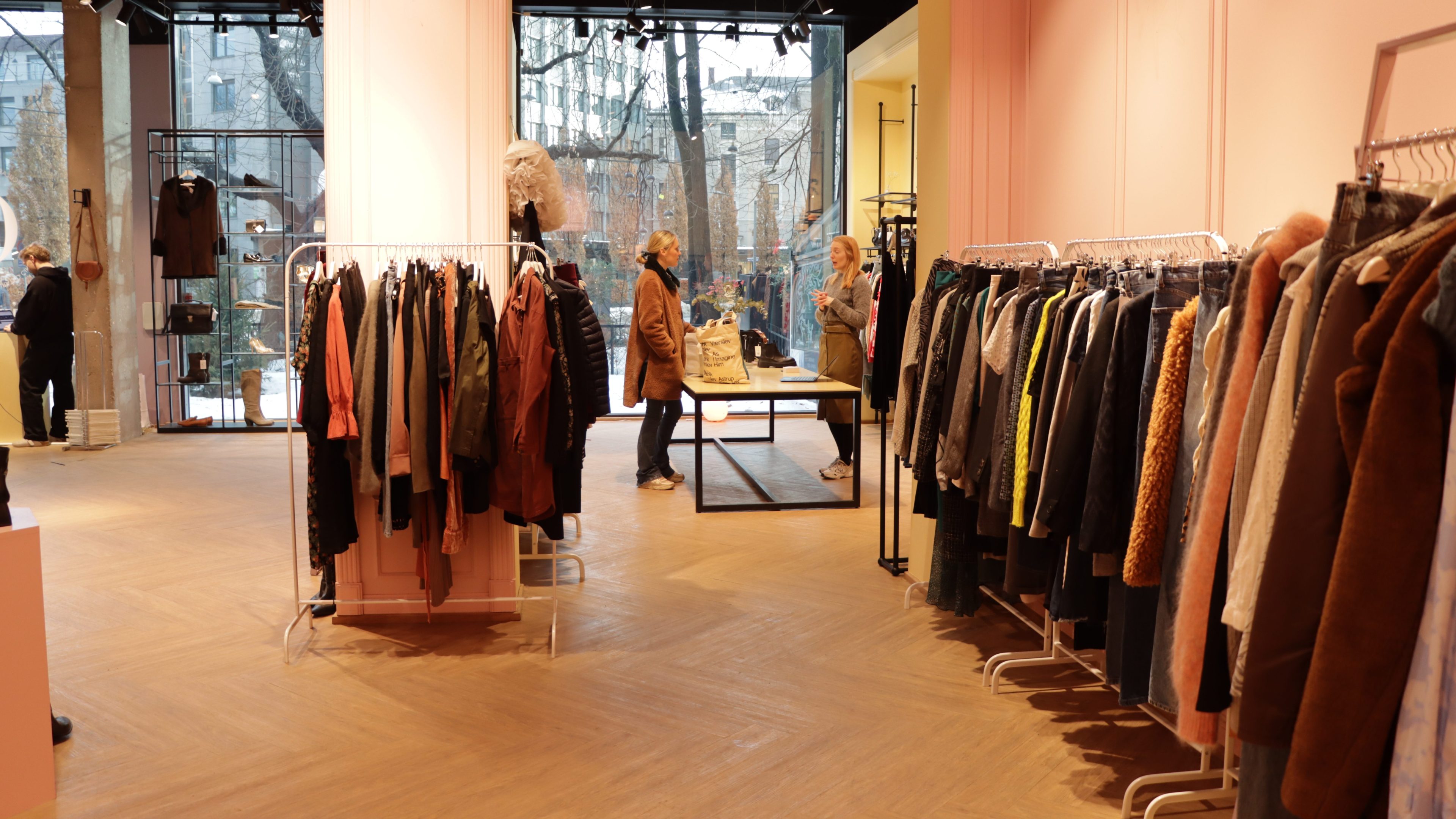 En stilig klesbutikk har stativer fulle av plagg, en elegant utstillingsdukke og to gjester som prater ved disken. Store vinduer avslører livlig gateutsikt utenfor, som minner om trendy bruktbutikker.