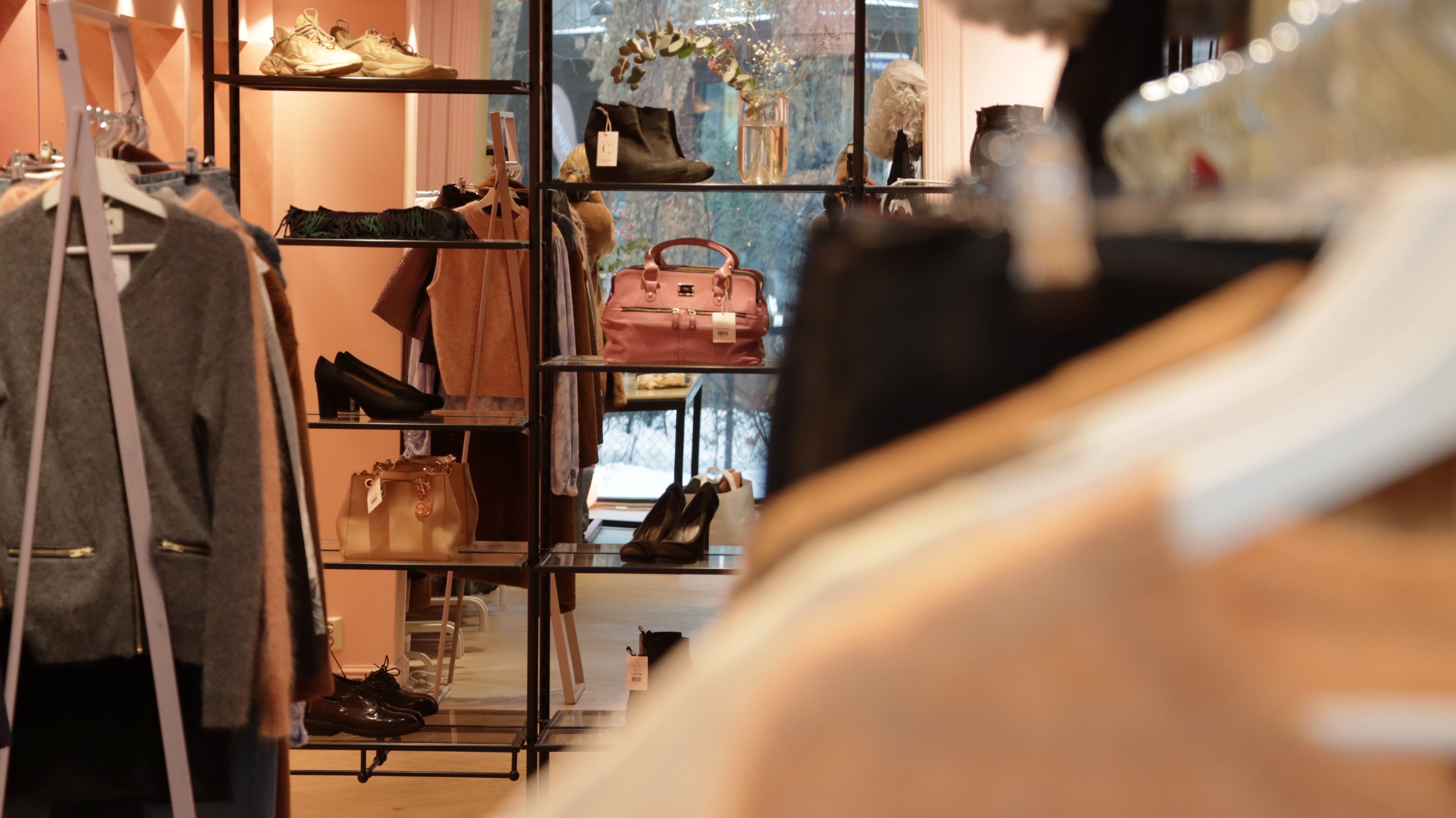 Interiøret i denne klesbutikken minner om sjarmerende bruktbutikker, med stativer med plagg og tilbehør smakfullt arrangert. En rosa veske står i sentrum, under dempet belysning og mot varme tregulv, og skaper en innbydende atmosfære.