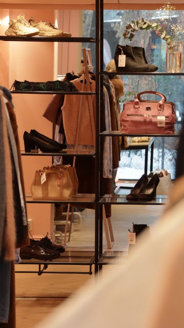 Butikkens interiør minner om elegante bruktbutikker, med hyller med sko, vesker og klær. Bakgrunnen har et vindu som viser en uskarp utendørsutsikt, og skaper en rolig shoppingstemning.