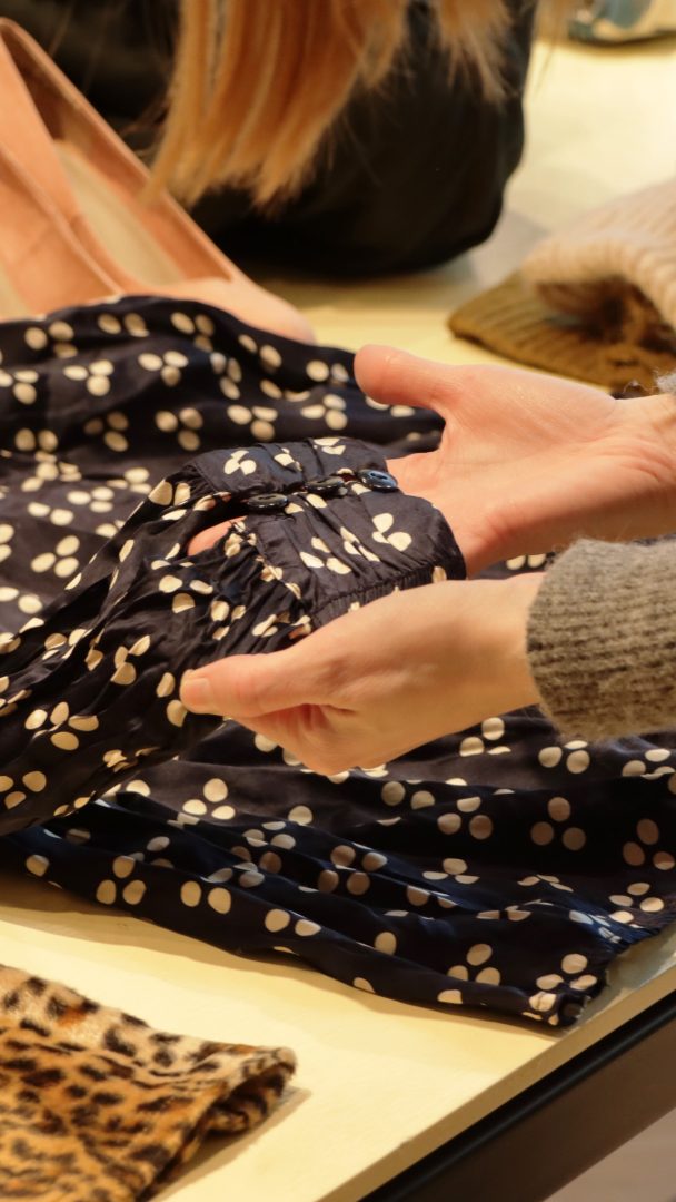 Hender undersøker nøye en marineblå kjole med hvite prikker lagt ut på et bord, sammen med et leopardmønster og en stilig solbrun bag, alle skatter fra lokale bruktbutikker.