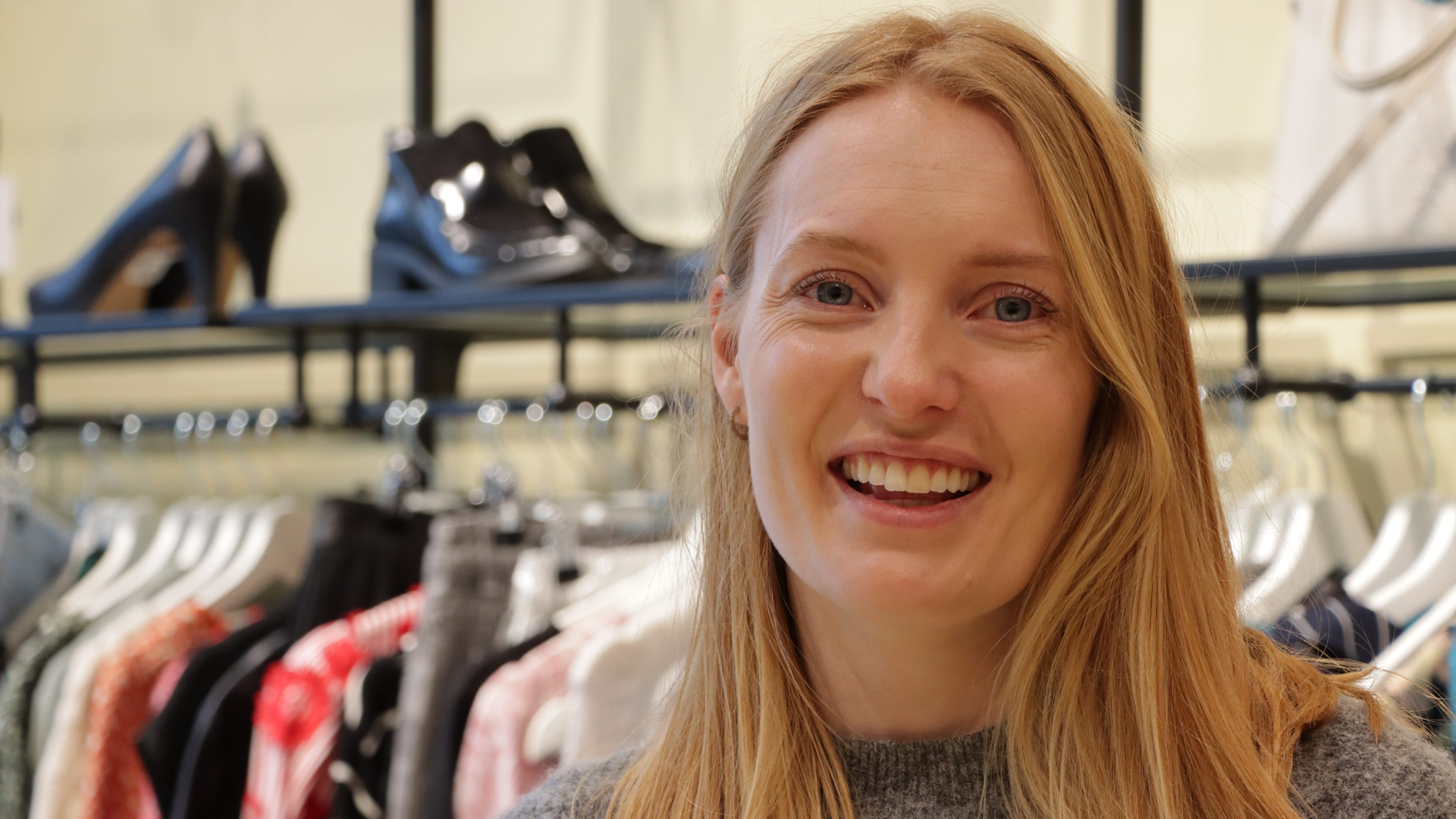 En kvinne med langt hår smiler i en klesbutikk, omgitt av stativer med klær og høyhælte sko. Gleden hennes gjenspeiler sjarmen som finnes i bruktbutikker, hvor hvert stykke forteller en unik historie som venter på å bli omfavnet.