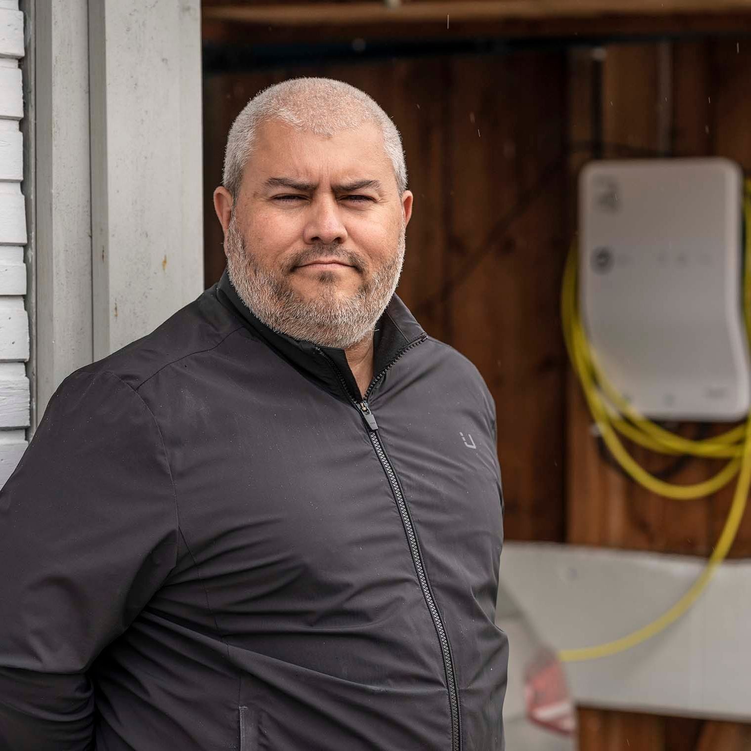 En mann med kort hår og skjegg står utenfor i mørk jakke. Bak ham ruver en trekonstruksjon mens gule kabler slanger seg langs veggen, og antyder nærliggende ladeplasser.
