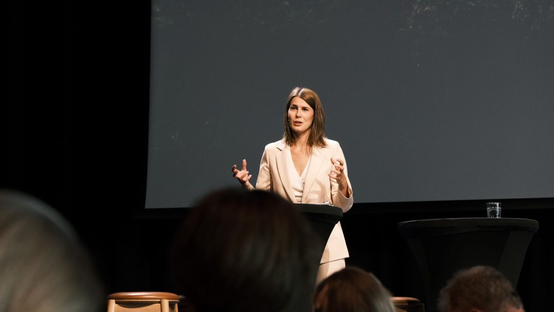 En kvinne i hvit dress snakker lidenskapelig ved et podium på en svakt opplyst scene.