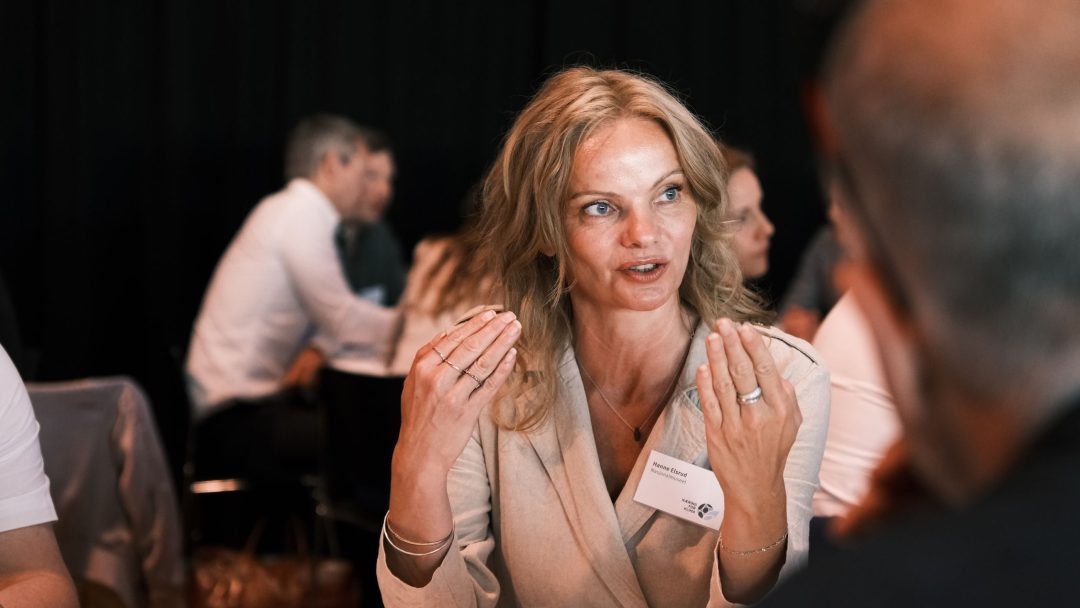 En kvinne i en lys blazer gestikulerer mens hun snakker på et møte, omgitt av andre deltakere. Hun har et navneskilt, og en kopp står på bordet foran henne.