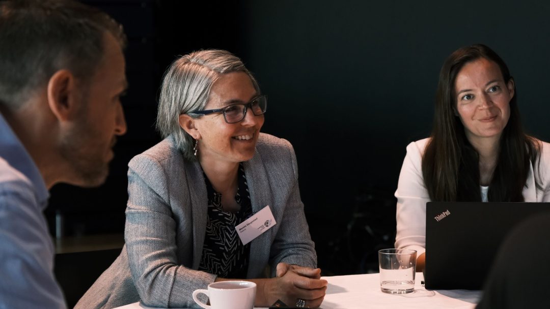 Tre personer som sitter ved et bord engasjert seg i en diskusjon; en har en bærbar datamaskin åpen. En kaffekopp og et glass vann står på bordet.