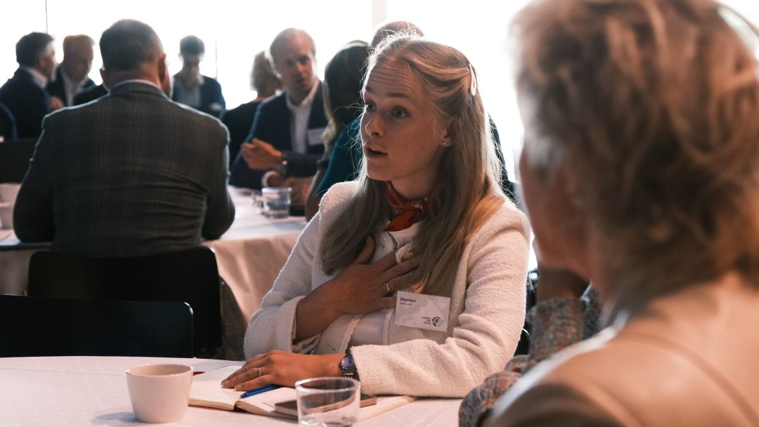 En kvinne med langt hår snakker ved en rundebordsdiskusjon, og holder den ene hånden mot brystet. Andre deltakere sitter rundt bordet.