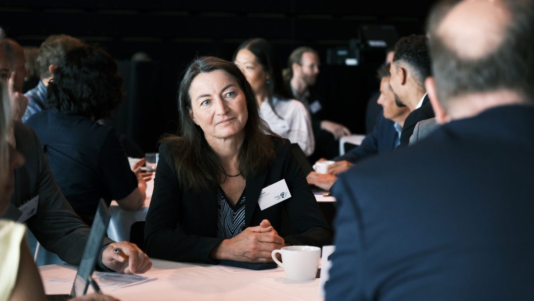 En kvinne sitter oppmerksomt ved et bord sammen med andre, engasjert i en diskusjon. Hun har navneskilt, og det står kopper og papirer på bordet.