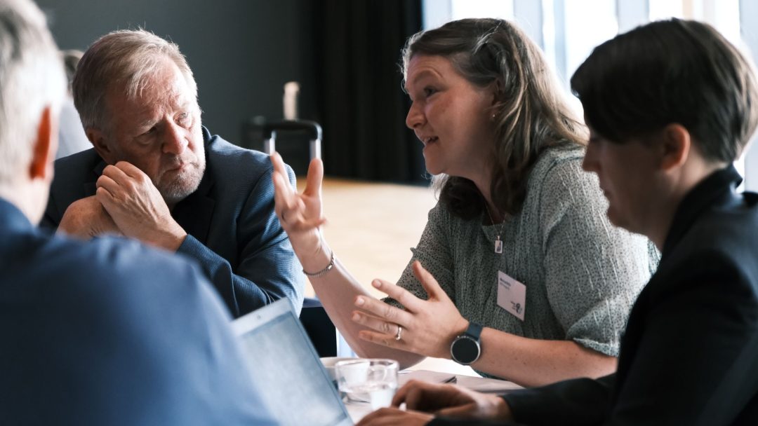 En gruppe mennesker engasjerte seg i diskusjon rundt et bord, med en kvinne som gestikulerte mens hun snakket.