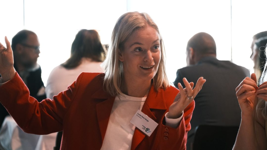 En person i rød frakk gestikulerer animert mens han snakker på en sammenkomst, med flere personer i bakgrunnen.