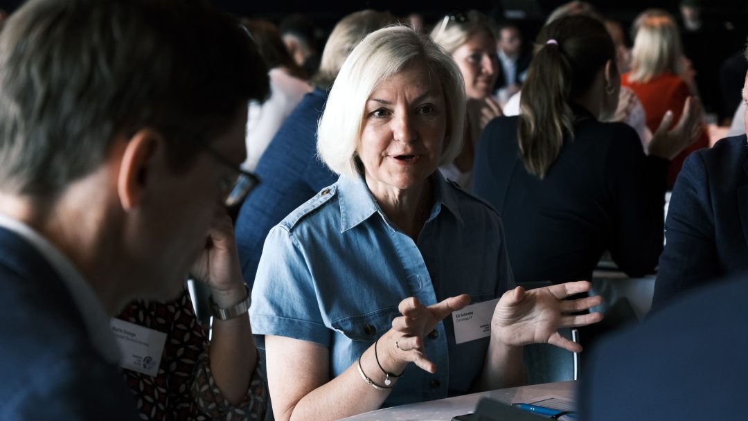 En eldre kvinne snakker livlig ved et rundt bord, omgitt av mennesker som er engasjert i diskusjon.