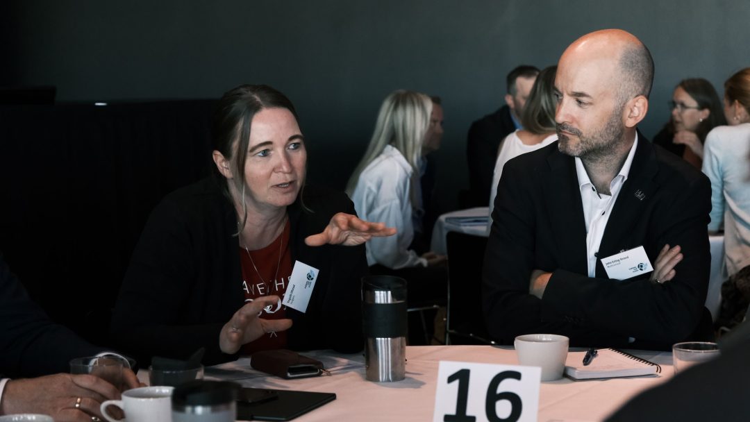 En kvinne og en mann i diskusjon ved et bord med kaffekopper og notatblokker, identifisert ved bord nummer 16. Flere personer er i bakgrunnen, også i samtale.