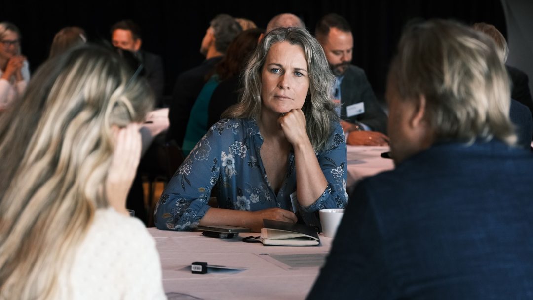 En kvinne sitter ved et bord med to andre under et møte eller en konferanse, og ser gjennomtenkt ut. Andre deltakere er synlige i bakgrunnen.