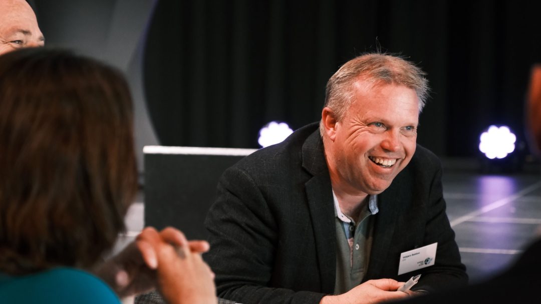 Mann som smiler og sitter ved et bord under et arrangement, med andre i nærheten.