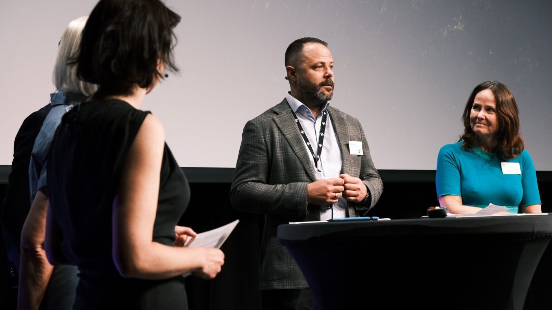 Fem personer deltok i en diskusjon på scenen, med én person som snakker mens andre lytter oppmerksomt.