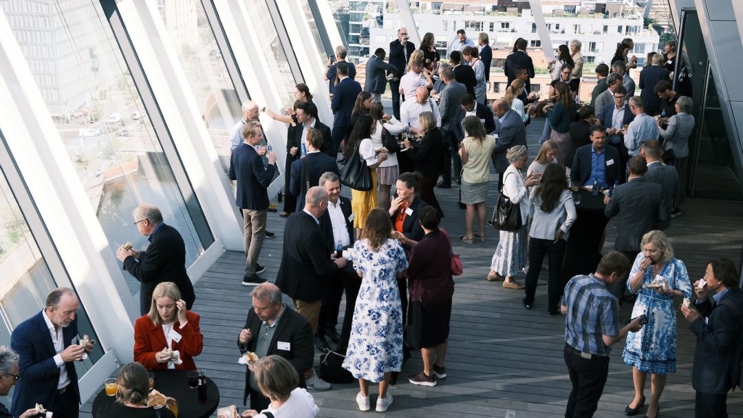 En stor gruppe mennesker mingler og sosialiserer seg på et formelt arrangement på en romslig, moderne terrasse med store vinduer og utsikt over byen.