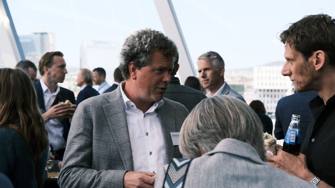 Folk engasjerte seg i samtale på et nettverksarrangement, med noen som holdt drinker. Lyse innendørs omgivelser med store vinduer og bybilde synlig i bakgrunnen.