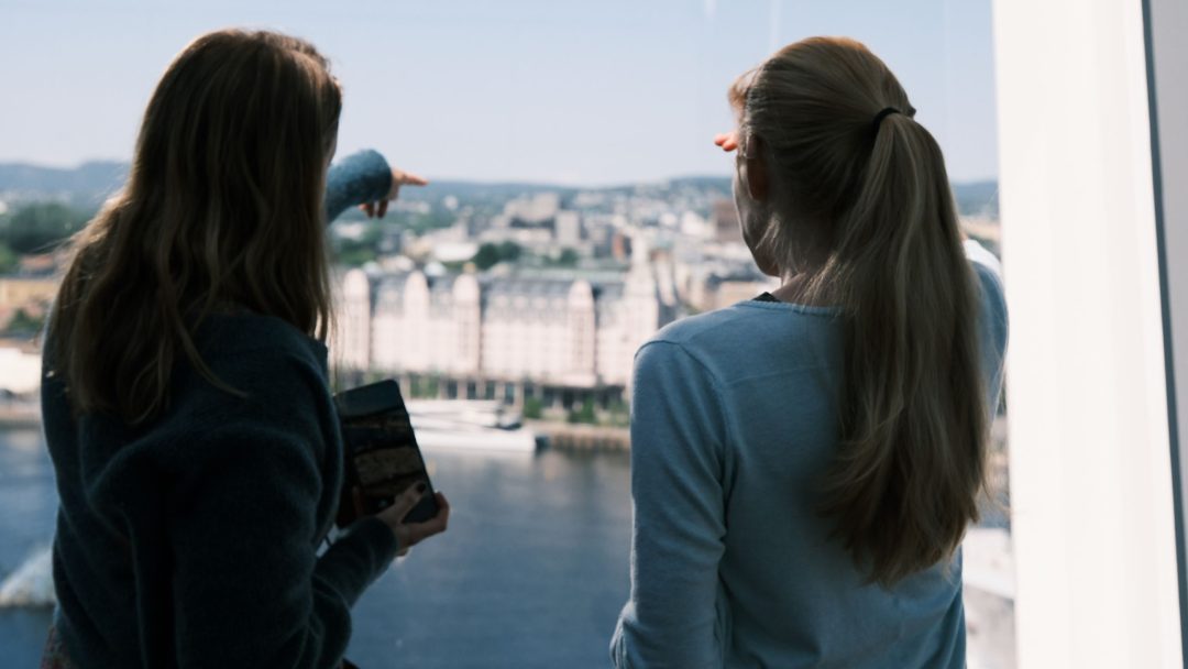 To kvinner står ved et vindu, med utsikt over en vannmasse og bybilde. Man peker utover mens man holder et nettbrett.