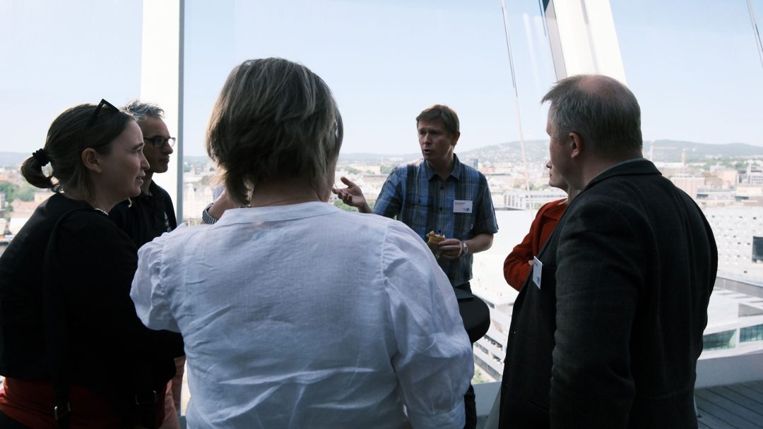 En gruppe mennesker står i samtale nær store vinduer med utsikt over et bybilde på en lys dag.