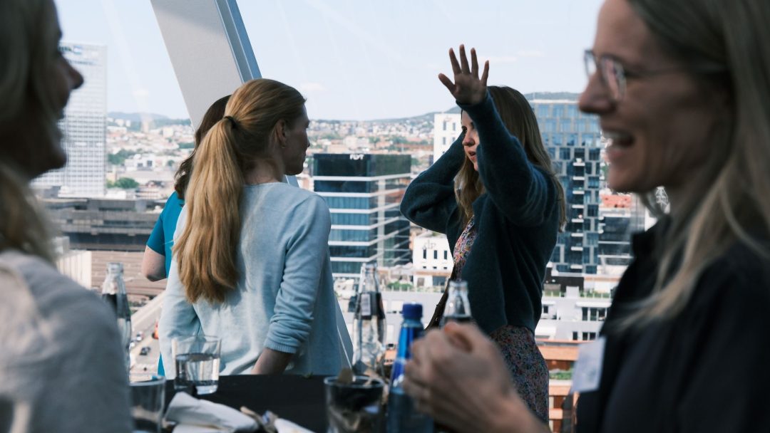 En gruppe mennesker snakker og ler på et høyhus med et bybilde i bakgrunnen. En person gestikulerer med løftede armer.