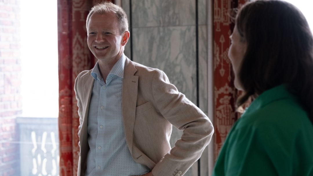 En mann i beige jakke og blå skjorte smiler mens han snakker med en kvinne i en grønn topp i et rom med mønstrede røde gardiner.