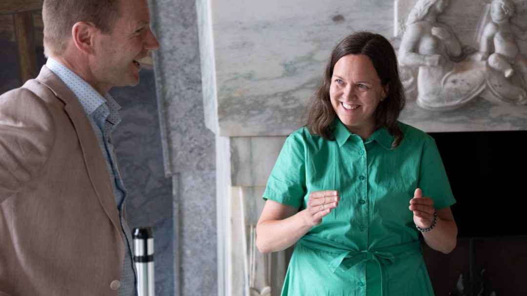 To personer er i samtale innendørs. Kvinnen i grønn kjole gestikulerer med hendene, mens mannen i solbrun jakke lytter. En dekorativ peis er i bakgrunnen.