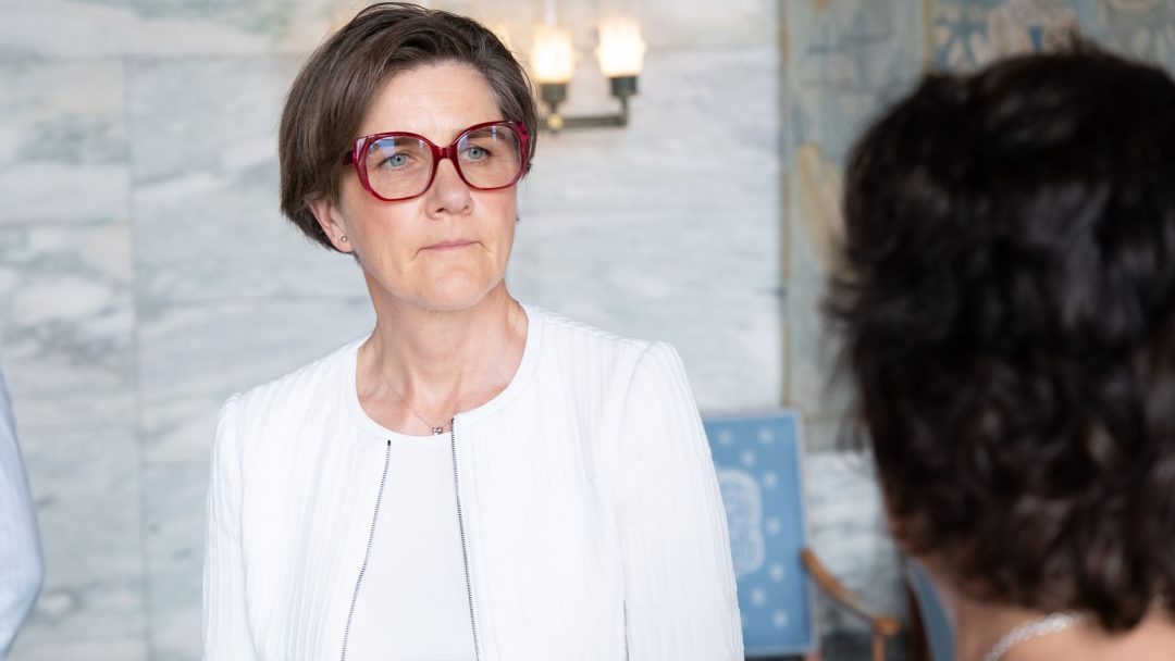 Person med store røde briller og hvit jakke, står innendørs og ser til siden med et gjennomtenkt uttrykk.