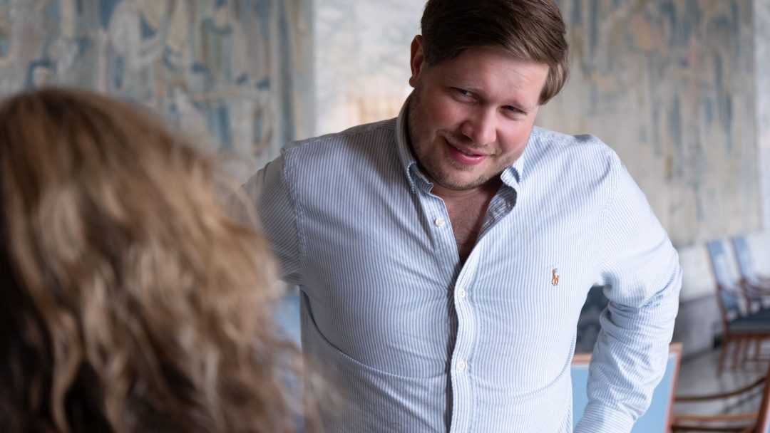 Mann i stripet skjorte med hendene på hoftene, lener seg fremover, snakker med en kvinne i et rom med mønstrede vegger.