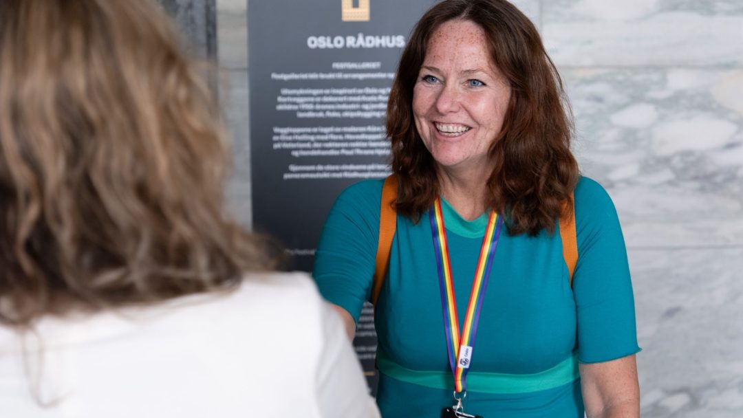 En kvinne i blågrønn kjole med regnbuesnor håndhilser på en annen person foran et «Oslo Rådhus»-skilt.