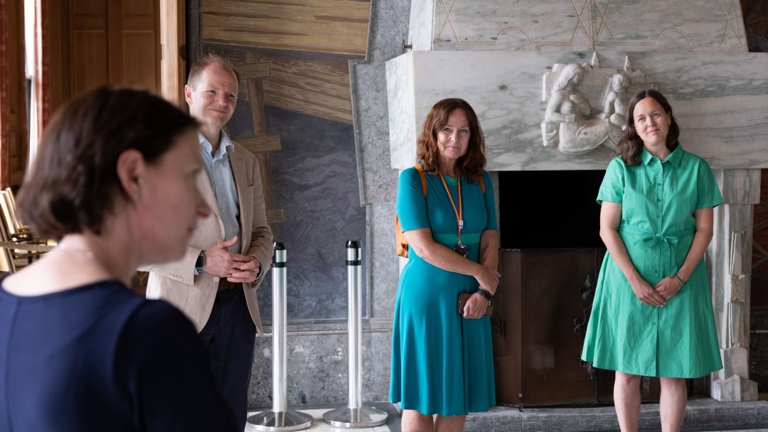 Fire personer står i et rom med en dekorativ steinpeis, vendt mot noen som snakker utenfor kameraet.